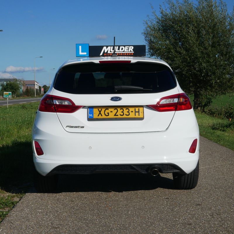 Achterkant van de witte Ford Fiesta lesauto met L-bord en Mulder-lesbord op het dak