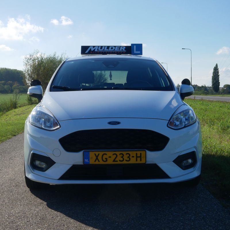 Voorkant van de witte Ford Fiesta lesauto met het Mulder-lesbord en L-bord op het dak